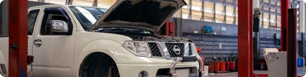 A white Nissan pickup truck with its hood open is parked inside an auto repair shop, surrounded by red vehicle lifts and equipment. auto repair services in South Jordan UT