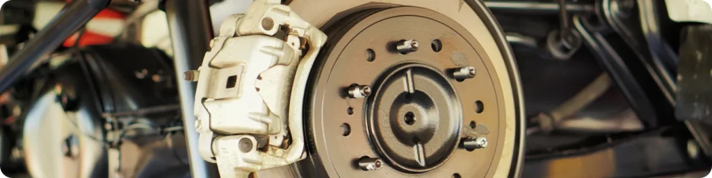 Close-up view of a car’s disc brake system, showing the brake caliper, rotor, and wheel studs on a vehicle with the wheel removed. The image focuses on the mechanical components and metal textures. brake repair, auto repair services in South Jordan UT