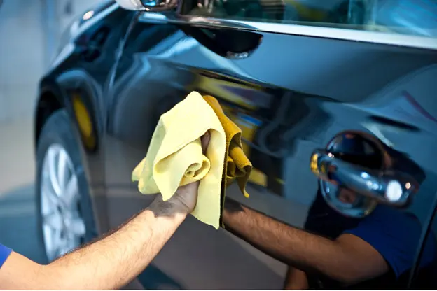 Car detailing in progress. Image shows a person using a yellow microfiber cloth to polish the exterior of a black car, focusing on the driver-side door.
