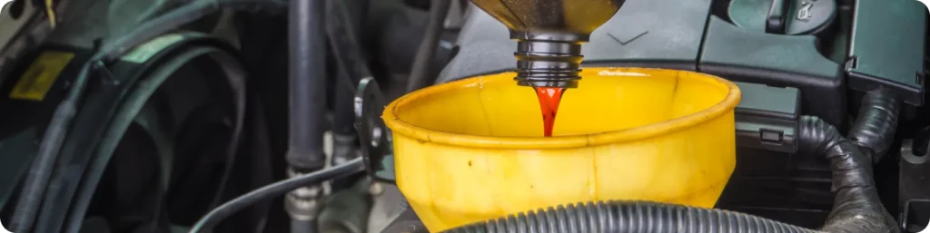 A close-up of red fluid being poured from a bottle into a yellow funnel placed in a car engine compartment, indicating an automotive fluid refill or maintenance process. Oil Change Services, auto repair services in South Jordan UT